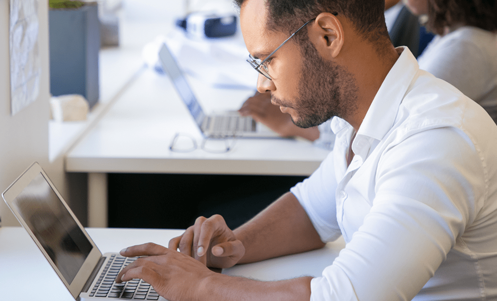 empresas. Homem de negro, usando óculos e uma camisa branca de manga longa, digitando em um laptop prateado sobre uma mesa branca. No fundo, há outras pessoas trabalhando.