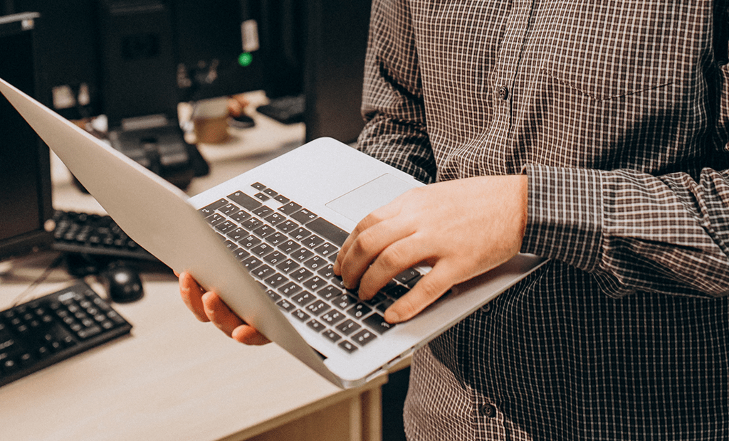 empresas. Pessoa vestindo uma camisa xadrez segurando um laptop aberto com uma mão e digitando com a outra. No fundo, há uma mesa com teclados e monitores.