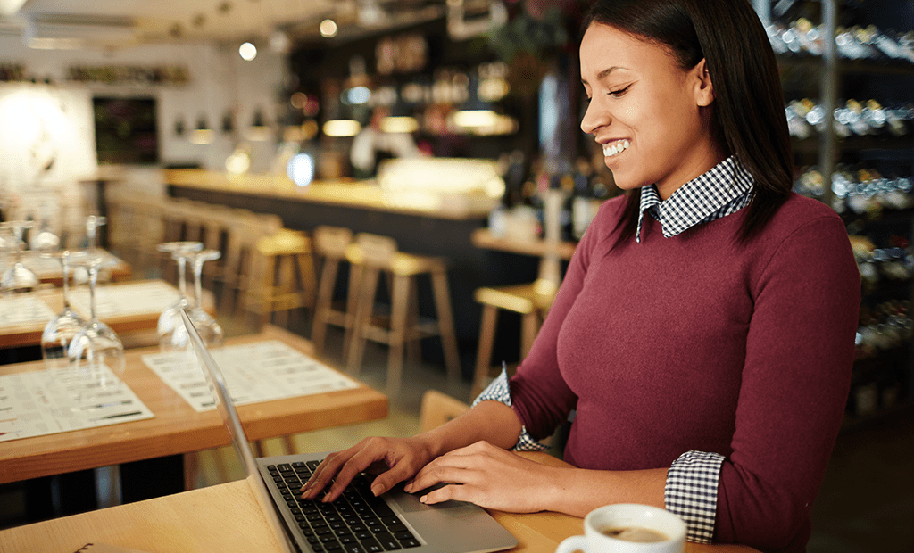 Acessibilidade Digital. Mulher sorridente trabalhando em um notebook dentro de um café. Ela está sentada à mesa, onde há uma xícara de café e cardápios. Ela veste um casaco vermelho e uma camisa xadrez por baixo.