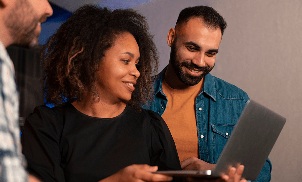 Acessibilidade digital. A imagem mostra três pessoas reunidas. Uma mulher de cabelo cacheado e roupa preta segura um tablet, enquanto um homem de barba, vestindo uma camisa jeans azul sobre uma camiseta marrom, olha para a tela de um notebook. Outro homem, veste uma camisa xadrez.