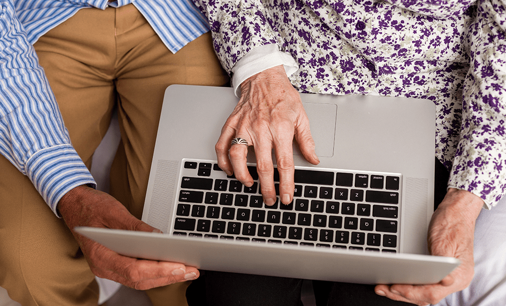 Acessibilidade digital. Duas pessoas idosas segurando um laptop. Uma delas veste uma camisa listrada azul e branca com calça bege, enquanto a outra usa uma blusa floral branca com roxo.