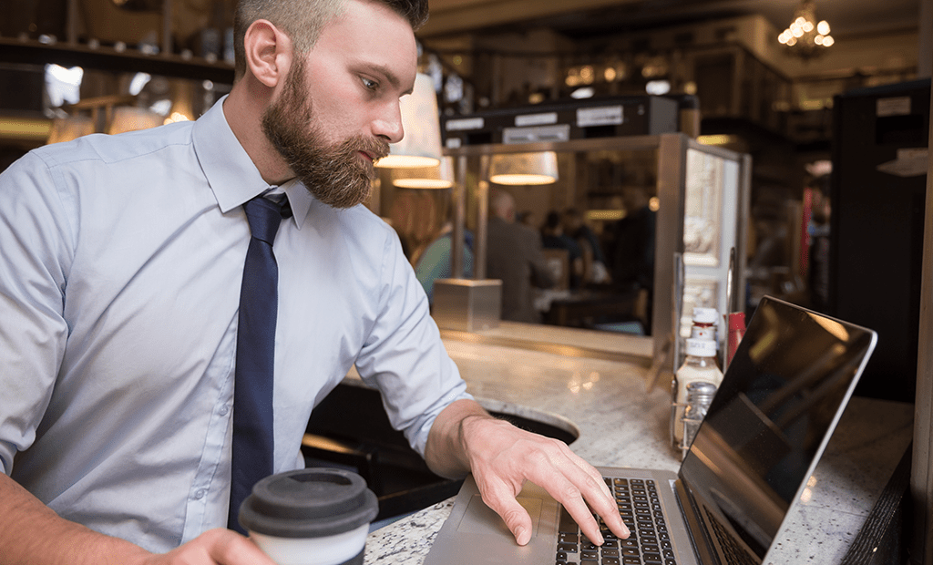 Acessibilidade Digital. homem de barba, vestindo uma camisa social azul clara e gravata azul escura, sentado em um balcão. Ele está digitando em um notebook enquanto segura um copo descartável de café.