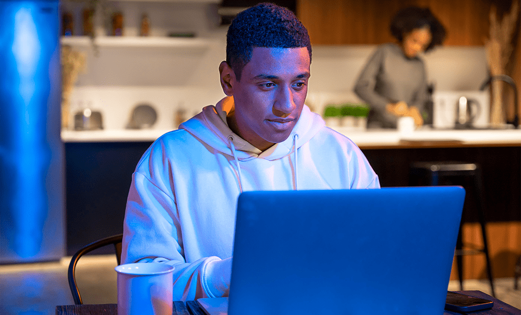 Autismo. Homem negro usando notebook em ambiente doméstico à noite, com xícara na mesa e pessoa ao fundo na cozinha.
