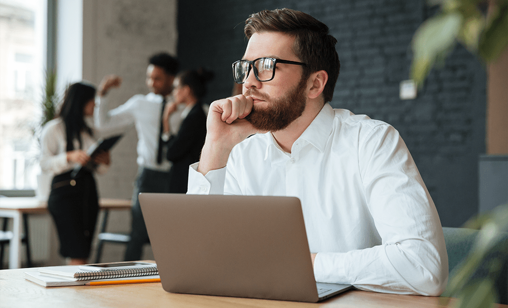 Overlays. Homem com barba, óculos e camisa branca, pensativo em frente a um notebook. Ao fundo, três pessoas conversam em pé.