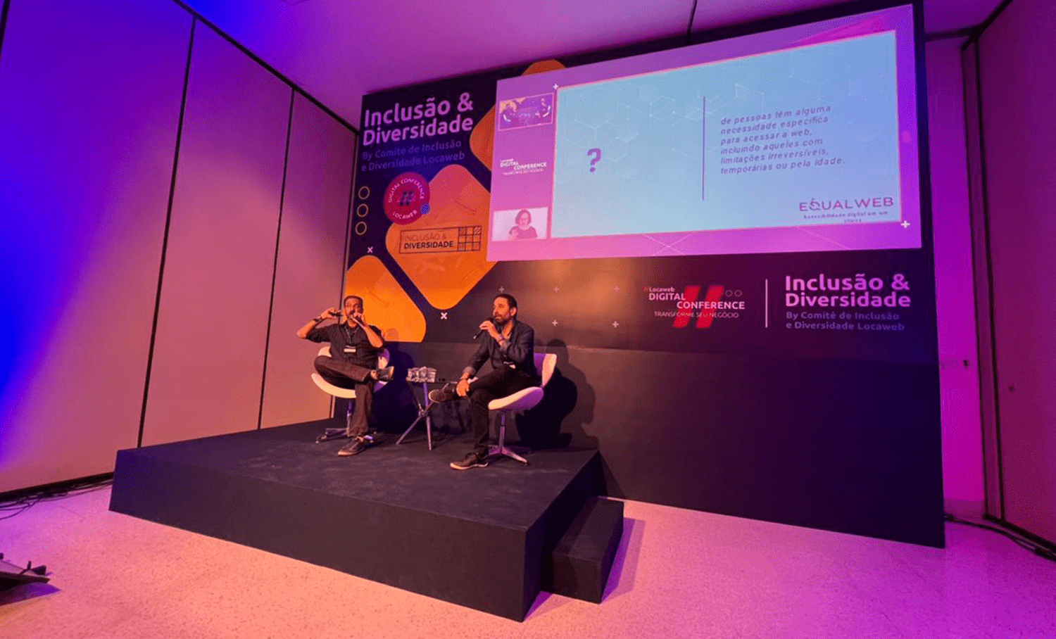 Dois palestrantes em palco com microfone. Painel exibe "Inclusão & Diversidade" e logo da EqualWeb na Locaweb Digital Conference.