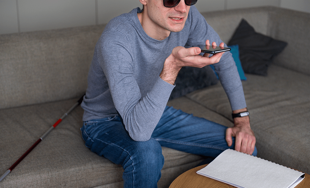 Homem com bengala e óculos escuros usa smartphone no modo áudio, com livro em braille à mão, ilustrando acessibilidade digital.
