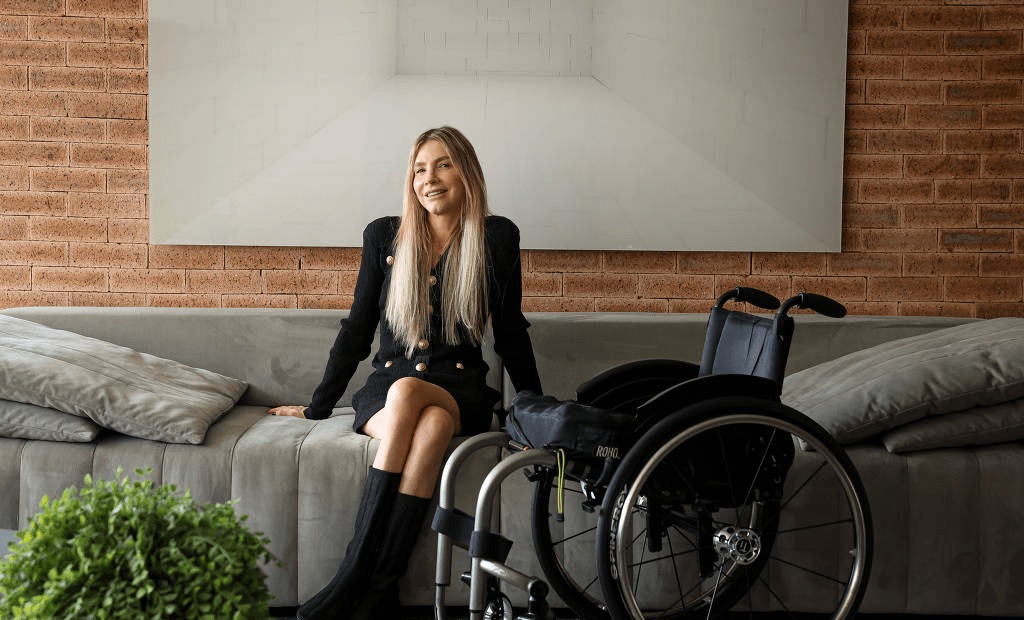 Andrea Schwarz sentada em sofá ao lado de sua cadeira de rodas, destacada na Folha de São Paulo por abordar o capacitismo na sociedade.