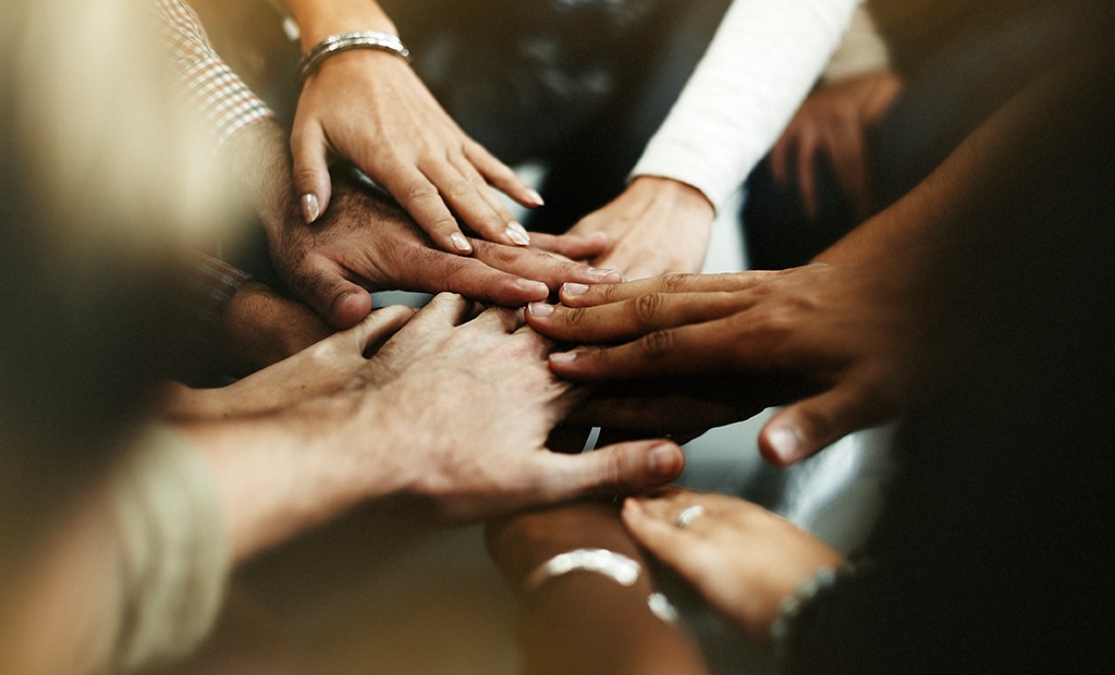 Múltiplas mãos de pessoas com diferentes tons de pele unidas no centro, simbolizando união, diversidade e colaboração em prol da inclusão.