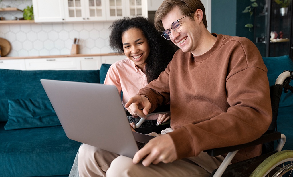 Homem em cadeira de rodas e mulher sorrindo enquanto usam um laptop juntos no sofá, ilustrando acesso tecnológico e inclusão digital.