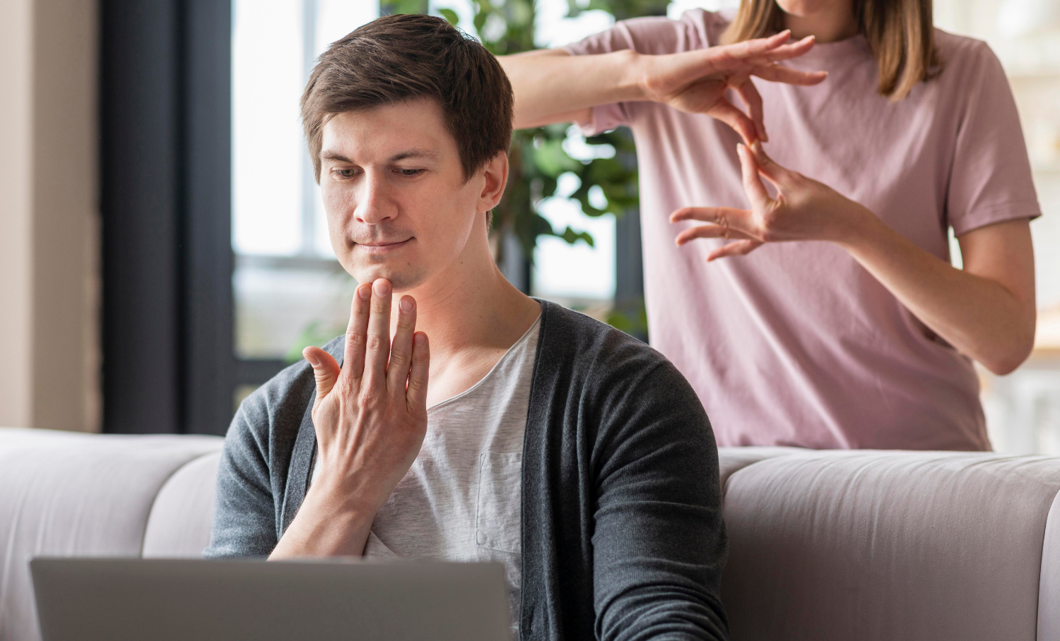 Homem usando laptop enquanto observa mulher fazendo LIBRAS ao fundo, ilustrando comunicação acessível para pessoas surdas.