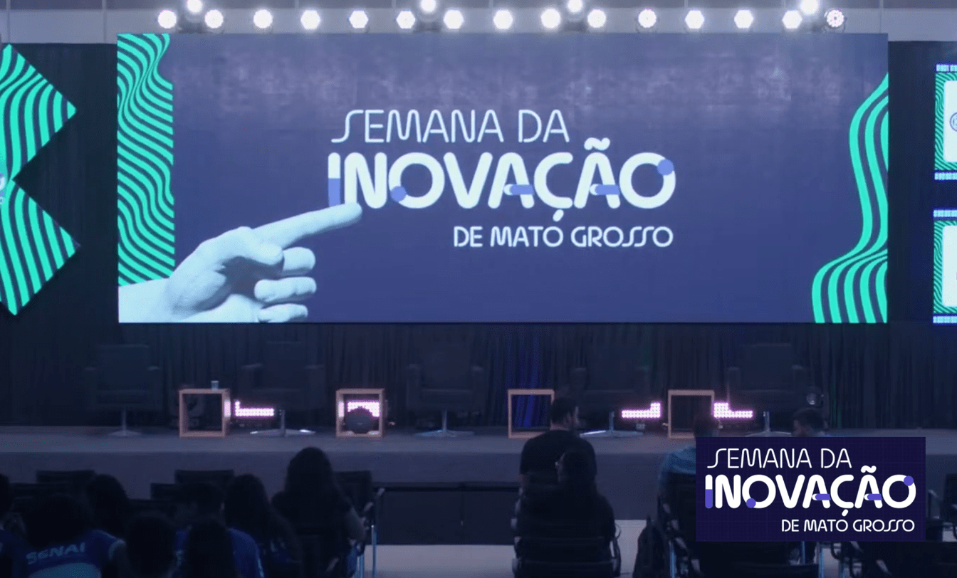 Palco da Semana da Inovação de MT com tela exibindo o logo e uma mão apontando. Plateia observa a área do debate sobre inclusão.