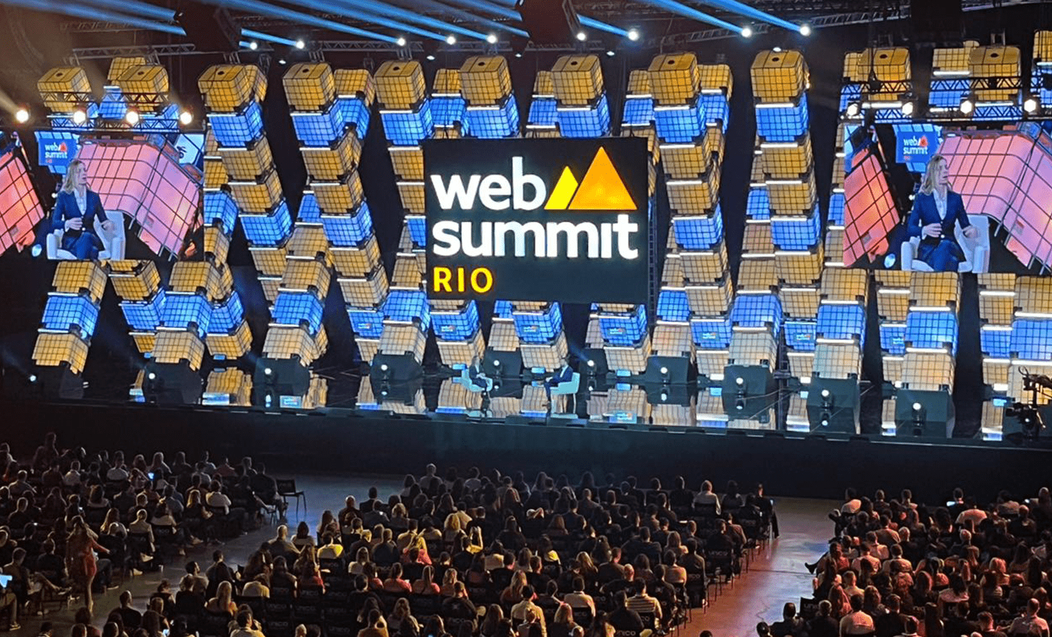 Palco do Web Summit Rio com telas exibindo o logo e palestrante, diante de grande plateia. Estrutura decorativa em cubos azuis e amarelos.