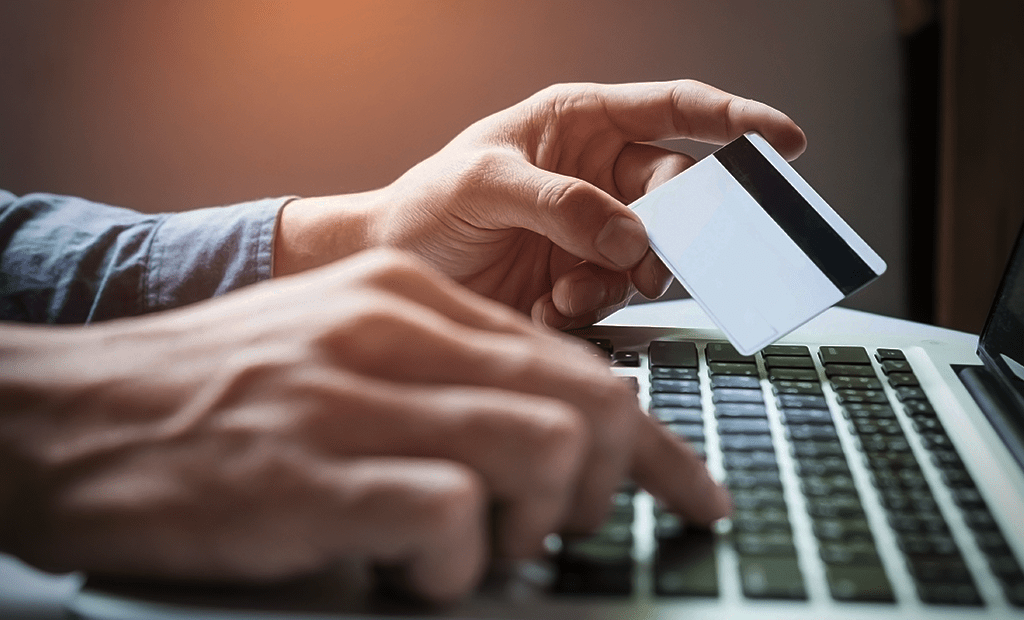 Mãos segurando um cartão branco com faixa magnética sobre o teclado de um laptop, simbolizando transações financeiras online.