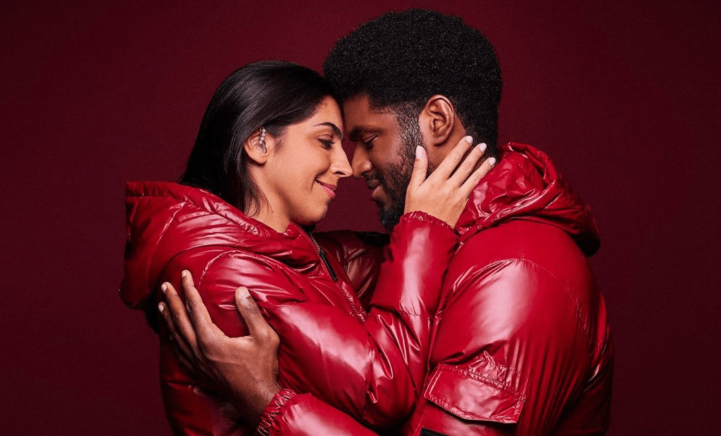 Casal se abraça com jaquetas vermelhas da Reserva; a mulher usa audifono. Cena promove amor e inclusão na campanha de Dia dos Namorados.