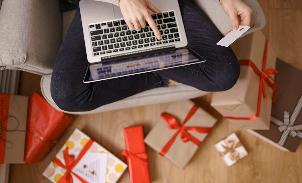 Pessoa compra online com laptop e cartão, cercada de presentes. Ilustra inclusão no comércio durante Black Friday e Natal.