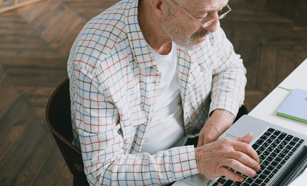 Homem mais velho com óculos e jaqueta xadrez digitando em laptop prateado sobre mesa branca, com caderno azul ao lado.