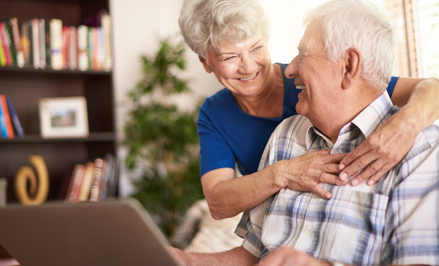 Casal de idosos sorrindo enquanto usa laptop juntos, ilustrando acesso e aprendizado tecnológico em ambiente doméstico acolhedor.