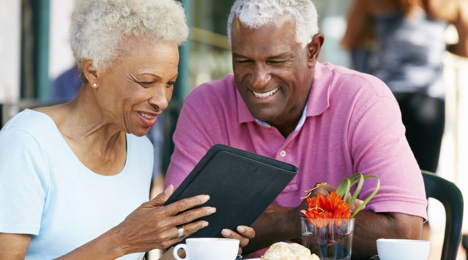 Casal de idosos sorrindo enquanto usa tablet em um café, ilustrando a acessibilidade digital e o engajamento do público +60 com tecnologia.