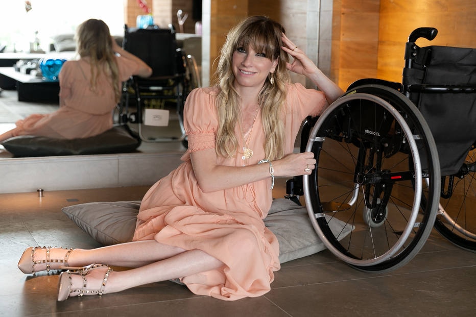 Mulher loira de vestido rosa sentada no chão ao lado de uma cadeira de rodas, sorrindo para a câmera com um espelho ao fundo.
