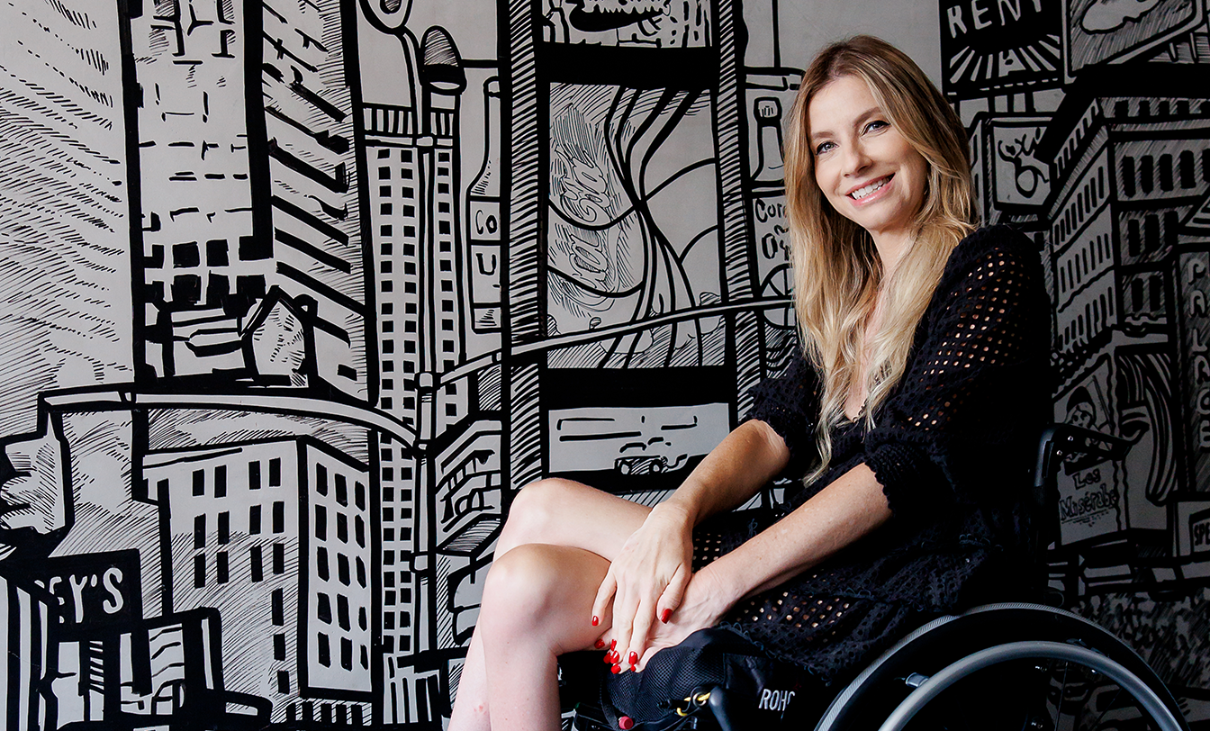 Mulher loira em cadeira de rodas sorrindo diante de mural urbano em preto e branco, ilustrando acessibilidade e inclusão social.