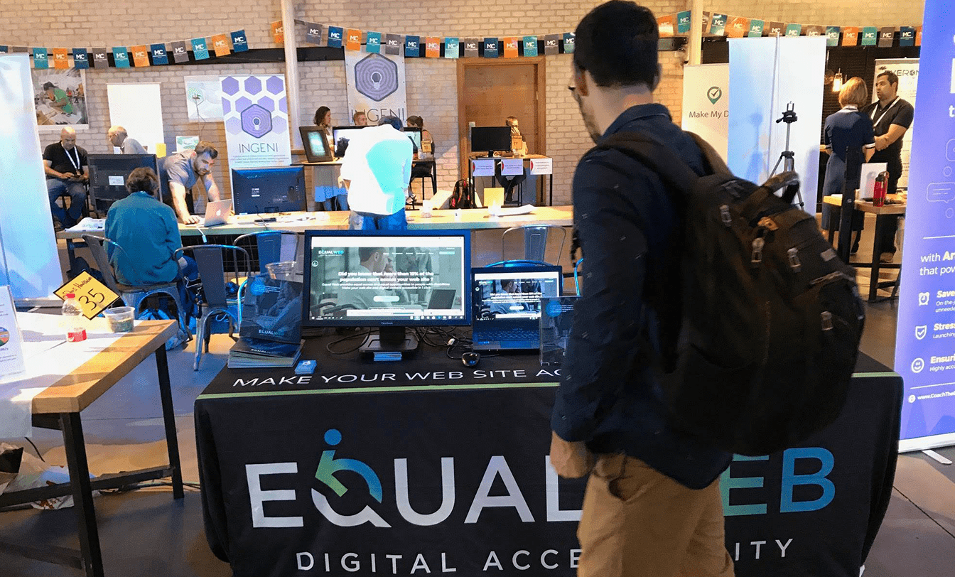 Stand da EqualWeb com mesa preta, logo e frase "Digital Accessibility". Monitores demonstram conteúdo de acessibilidade.