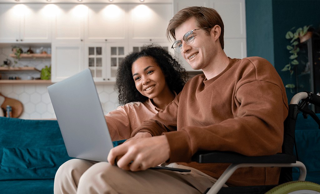 Homem em cadeira de rodas e mulher usam laptop no sofá, ilustrando inclusão digital e empoderamento via acessibilidade tecnológica.