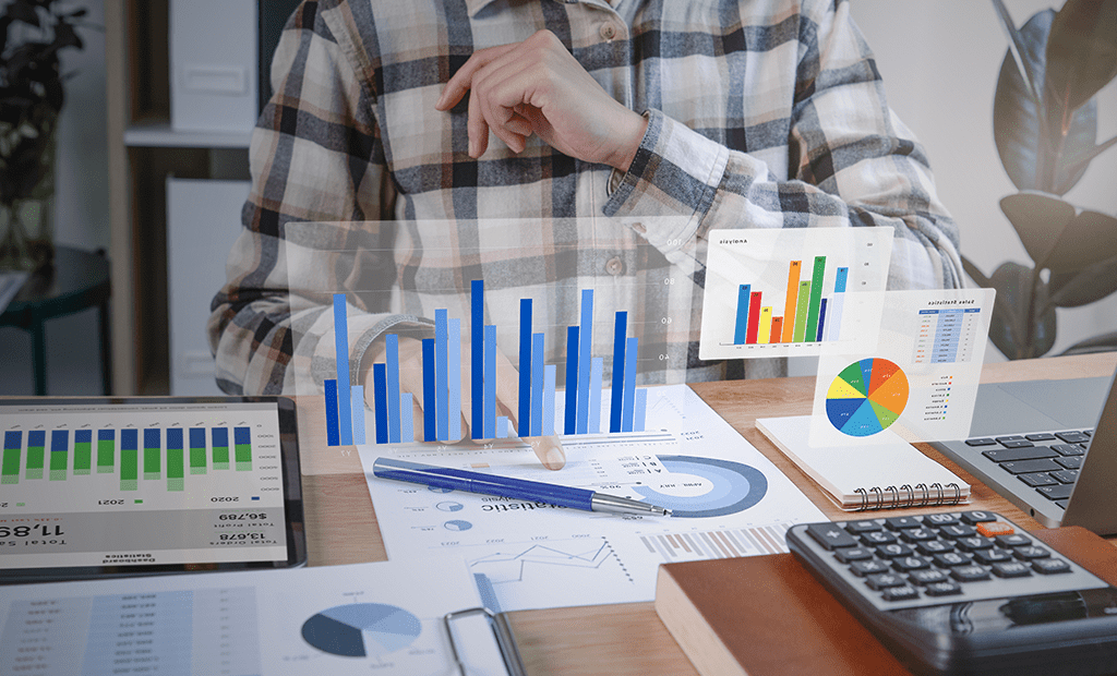 ROI. Uma pessoa, vestindo uma camisa xadrez casual, analisando dados financeiros em um ambiente de escritório. Há gráficos coloridos de barras, pizza e outros dados exibidos tanto em papel quanto em dispositivos digitais.