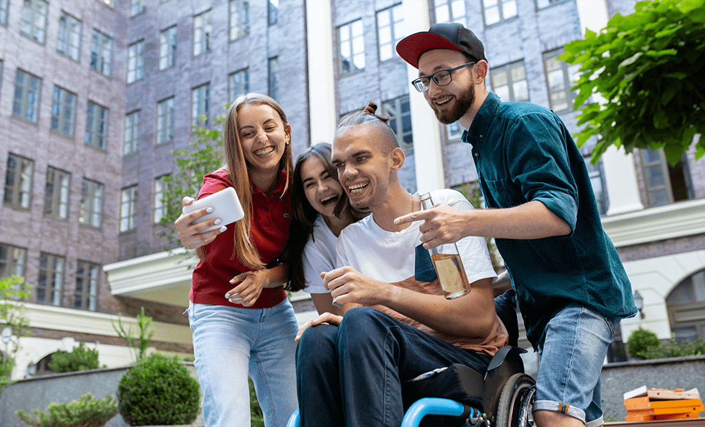 Jovem em cadeira de rodas tira selfie com amigos sorrindo ao ar livre, ilustrando inclusão e acessibilidade em eventos sociais.