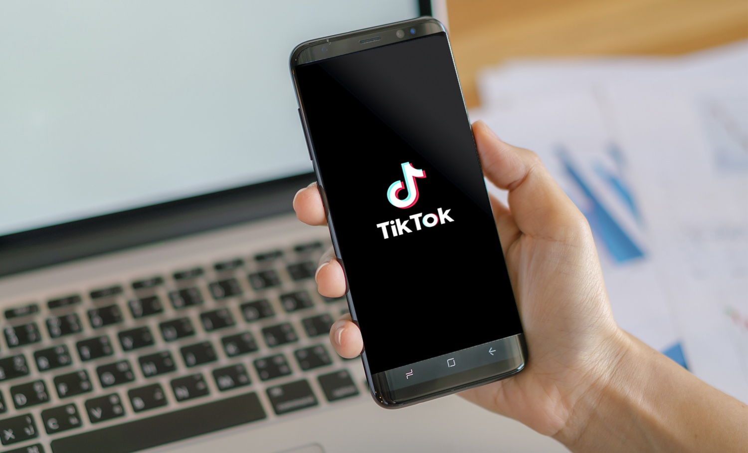 Mão segurando smartphone com o logo do TikTok na tela, sobre fundo com laptop e documentos.