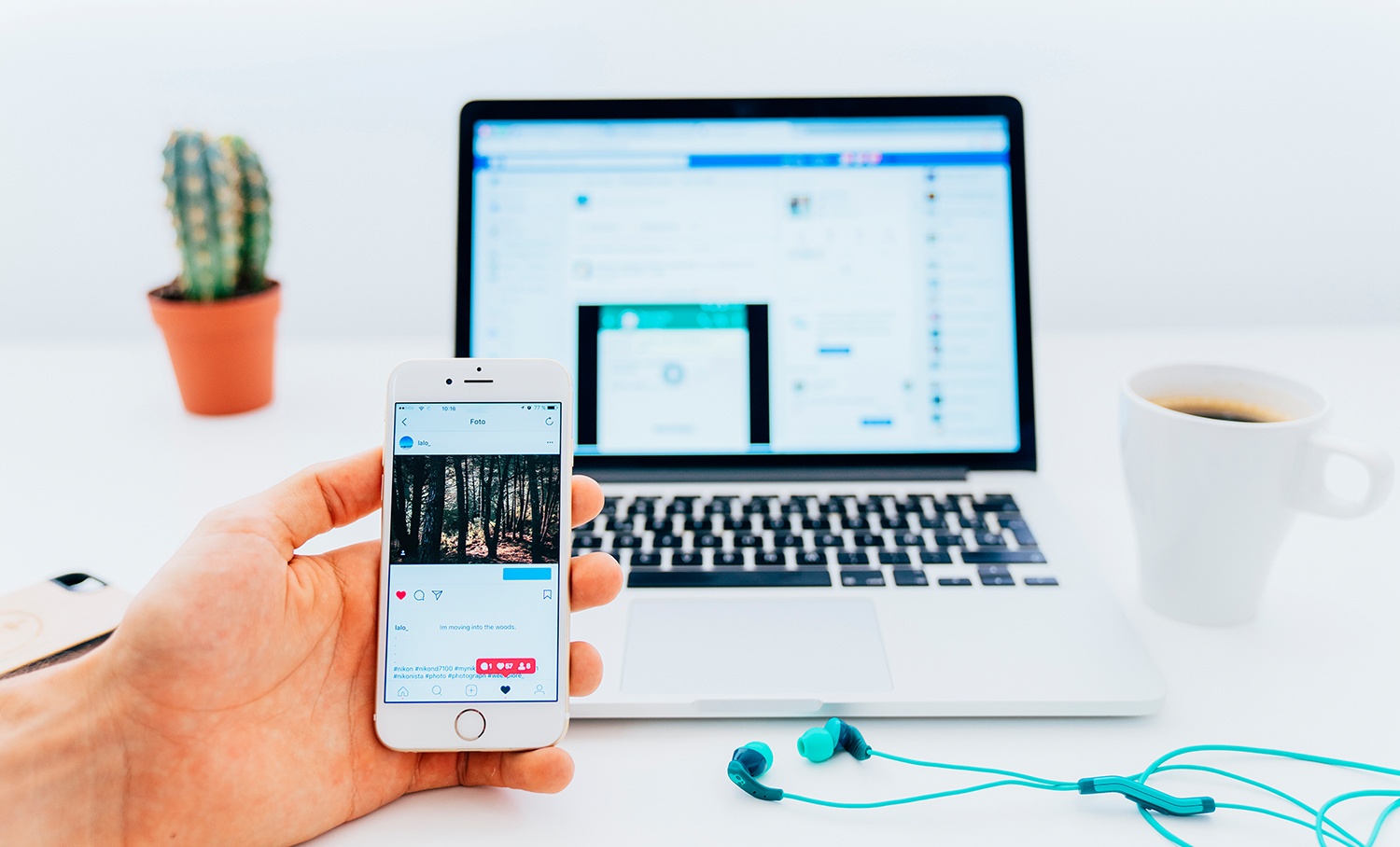 Mão segurando smartphone com post de rede social, laptop ao fundo e fones de ouvido na mesa branca.