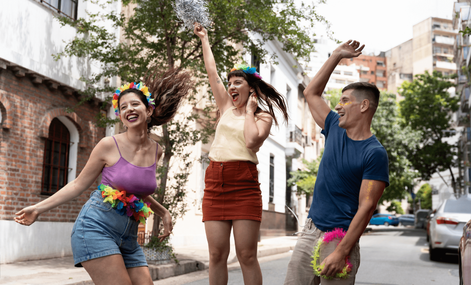Três jovens dançam e celebram em uma rua urbana, usando coroas coloridas e adereços festivos, representando a alegria do Carnaval nas ruas.
