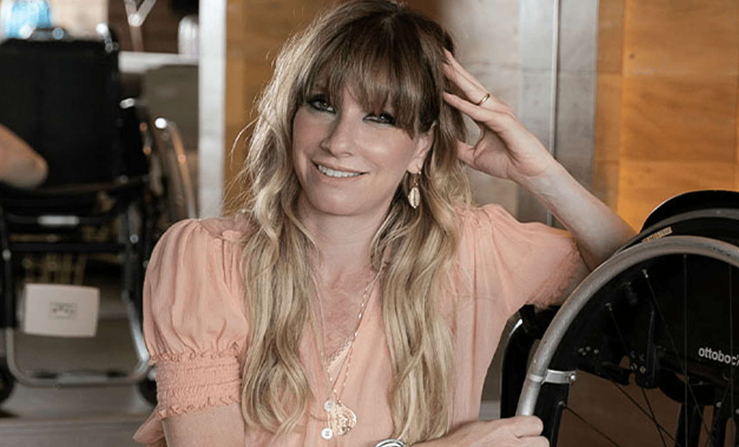 Andrea Schwarz sorrindo, com cabelos loiros e blusa rosa, apoiada em uma cadeira de rodas Ottobock, em ambiente interno.