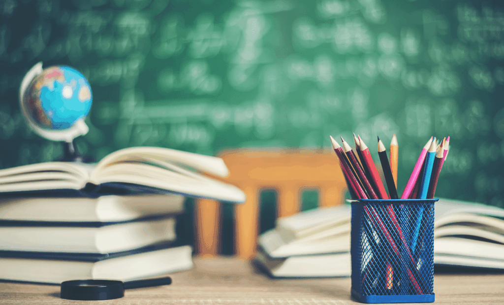educação. Imagem de uma mesa de estudo com alguns livros empilhados e outros abertos em cima da mesa. Do lado direito podemos observar um porta lápis com alguns lápis dentro e do lado esquerdo uma lupa.