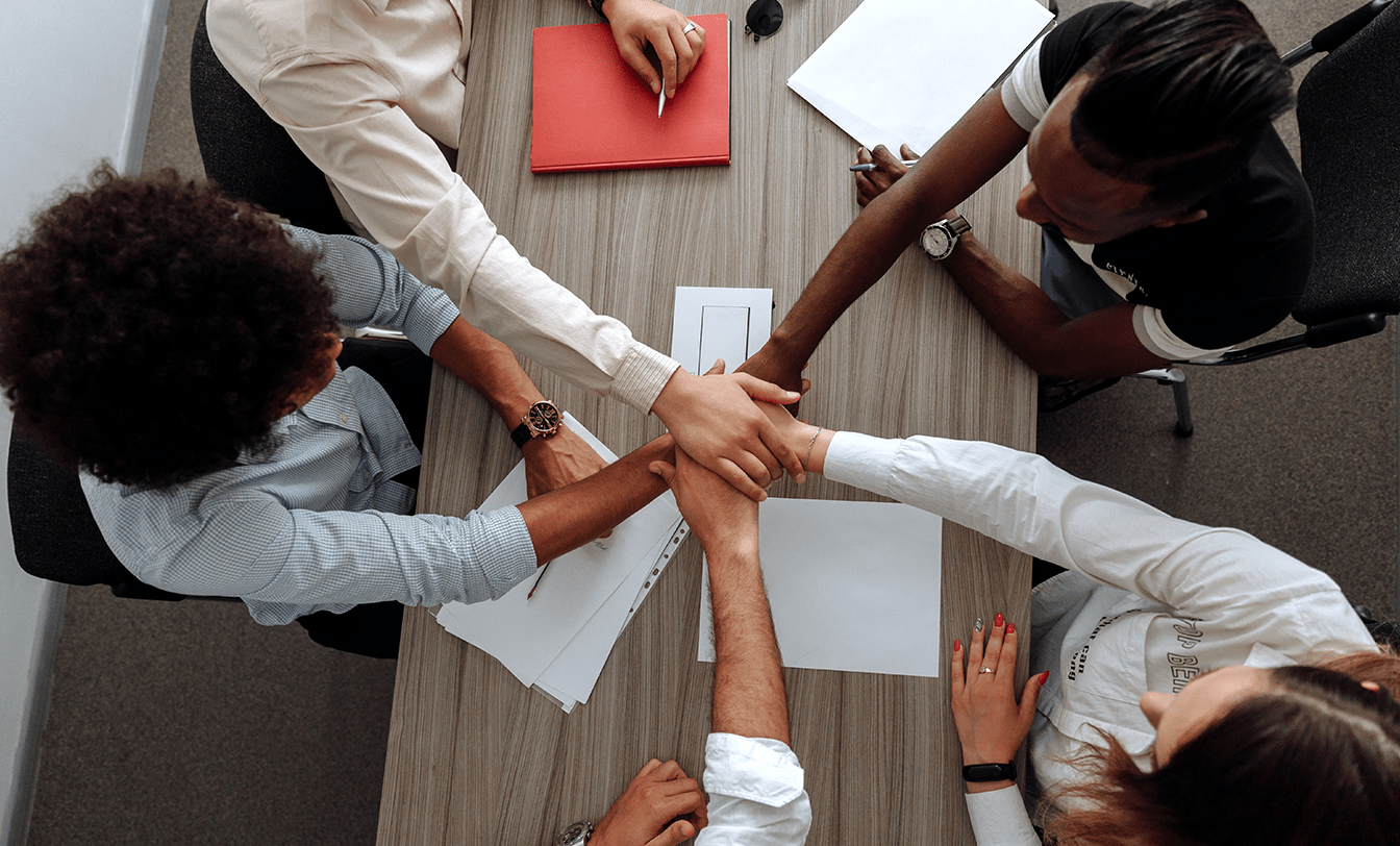 Vista aérea de mãos de pessoas diversas empilhadas sobre uma mesa de reunião, simbolizando união e trabalho em equipe corporativo.