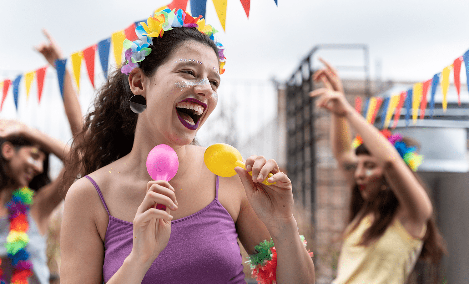 Mulher rindo com coroa de flores e maracas coloridas em festa com bandeirinhas, celebrando o Carnaval com alegria e inclusão.