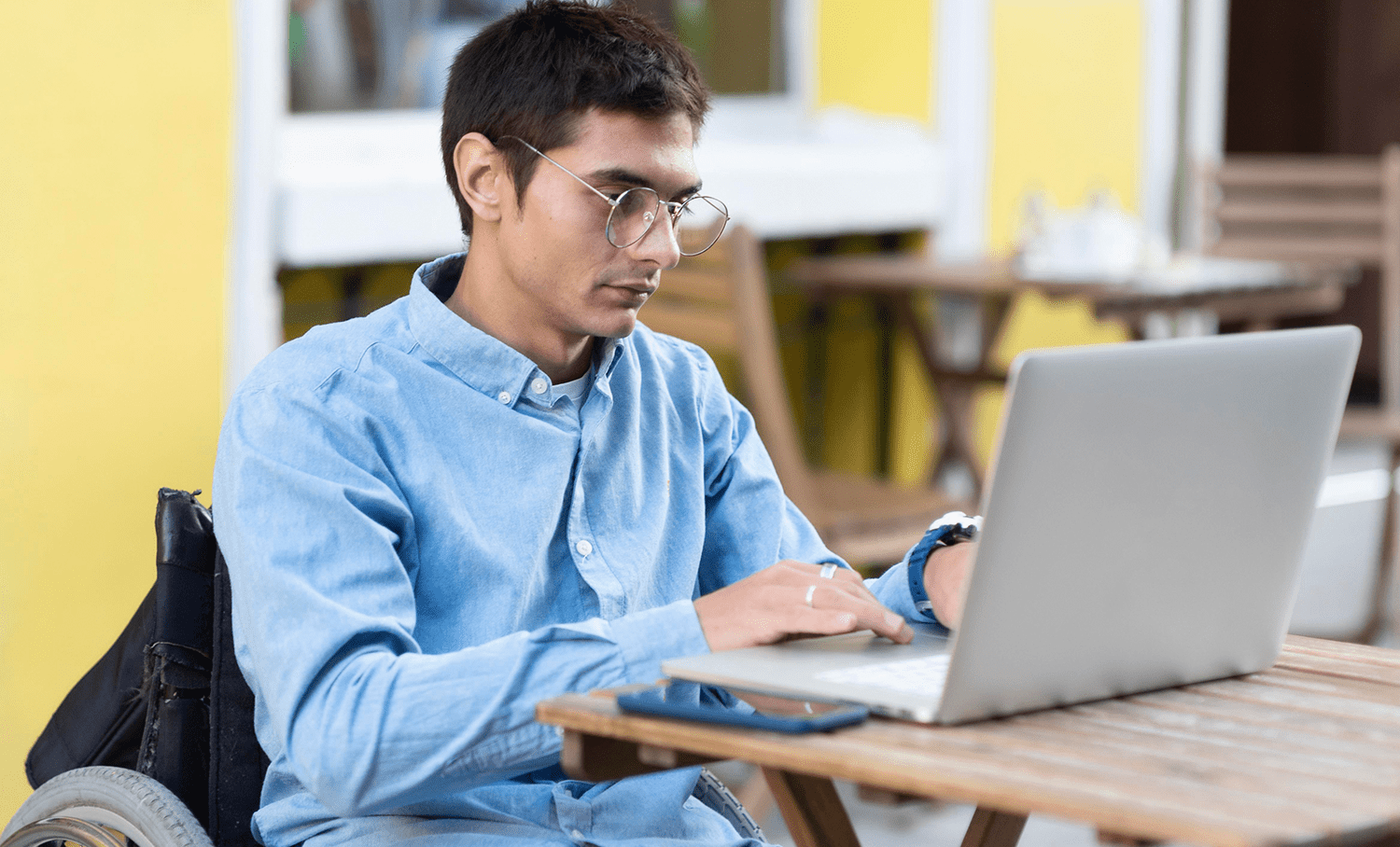 Homem em cadeira de rodas usando laptop em uma mesa ao ar livre, ilustrando acessibilidade e inclusão digital promovida pela EqualWeb.