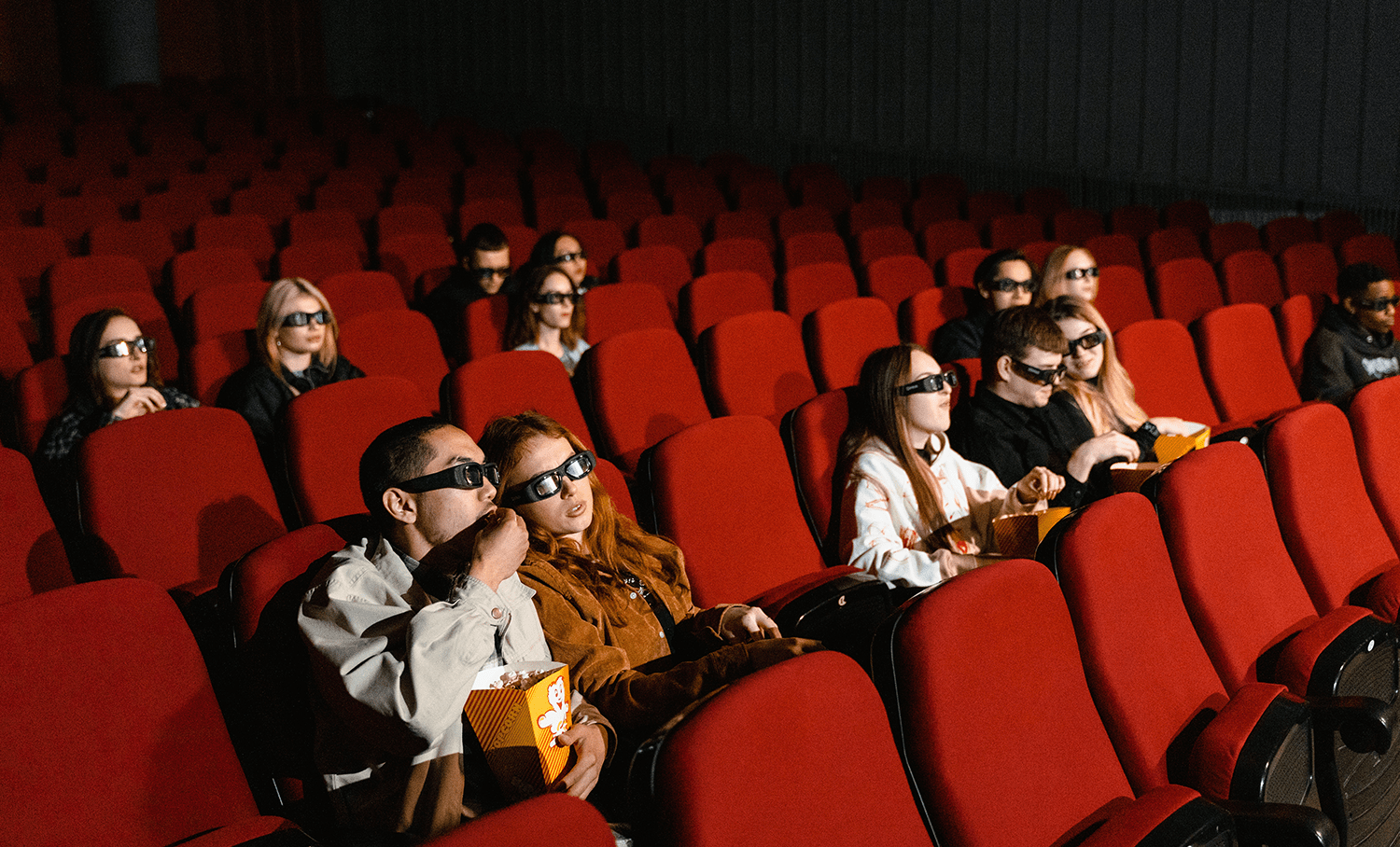 Público com óculos 3D assistindo a um filme em sala de cinema com poltronas vermelhas, ilustrando a experiência de entretenimento visual.