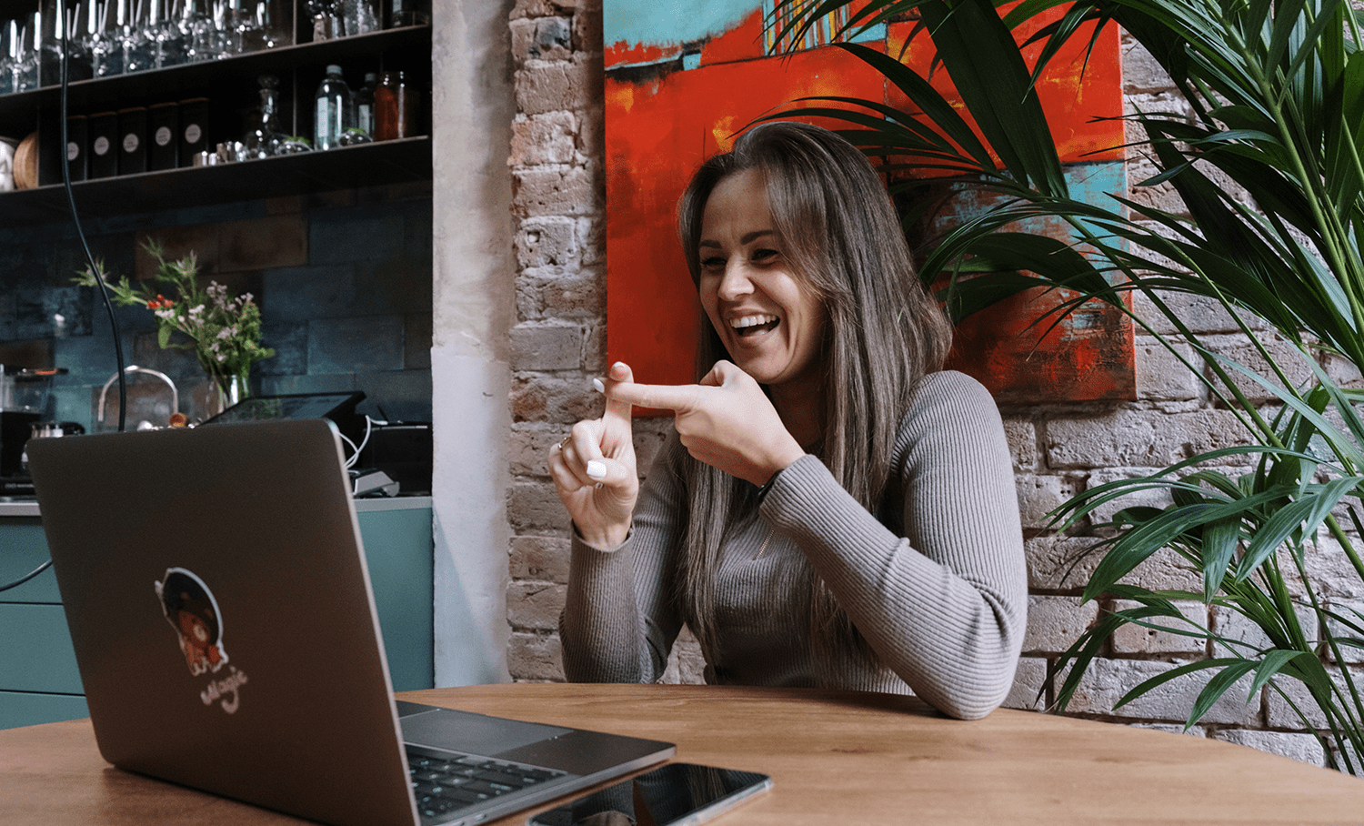 Mulher sorridente em videochamada no laptop, gesticulando com os dedos. Cena ilustra comunicação acessível e interação digital inclusiva.