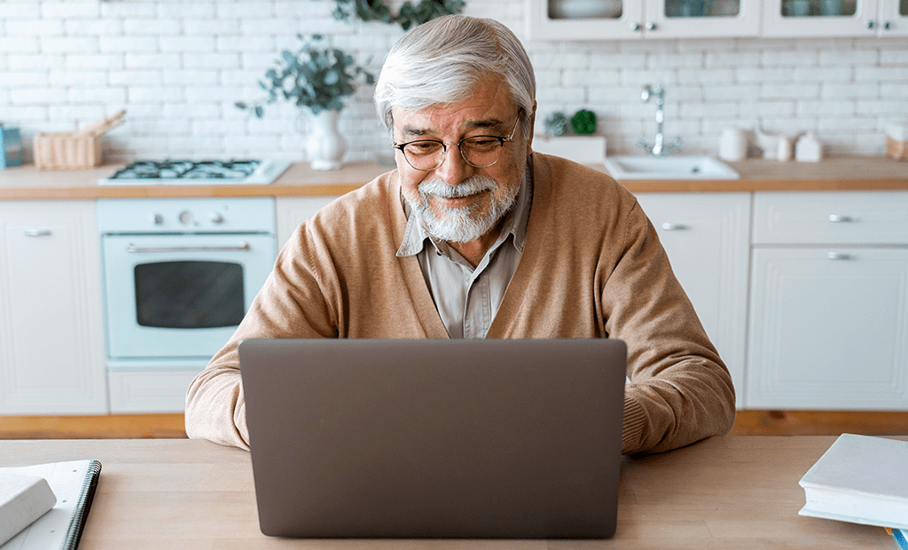 Homem mais velho com óculos usa laptop sorrindo em cozinha, ilustrando acesso digital inclusivo e autonomia na tecnologia.