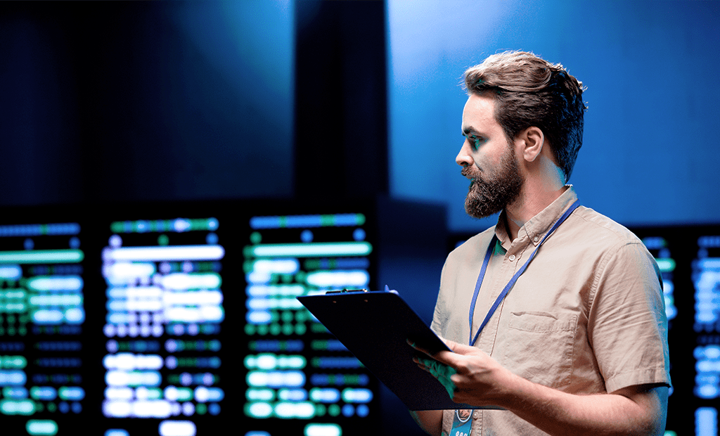 Homem com barba segura prancheta em frente a monitores com dados, representando análise tecnológica e inovação digital para acessibilidade.