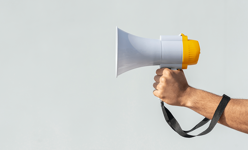 Mão segurando um megafone branco e amarelo com alça preta, simbolizando comunicação, voz e a necessidade de acesso universal à informação.