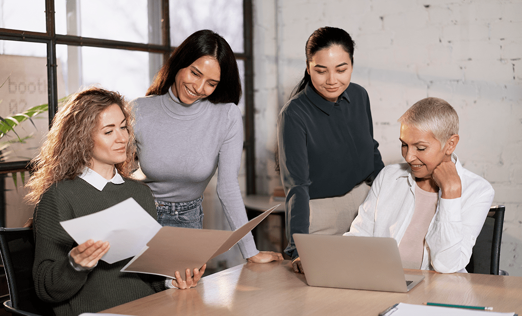 Quatro mulheres de diferentes gerações e etnias colaboram num escritório, representando diversidade e liderança feminina corporativa.
