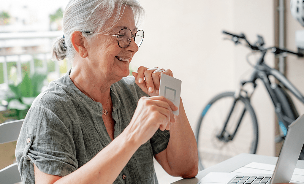 Mulher idosa sorridente segura cartão de crédito diante do laptop, ilustrando compra online acessível e inclusiva para o Dia do Consumidor.