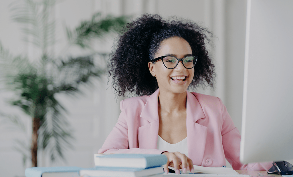 Mulher sorridente com óculos e blazer rosa working no computador, ilustrando produtividade e acessibilidade digital em ambiente de trabalho.