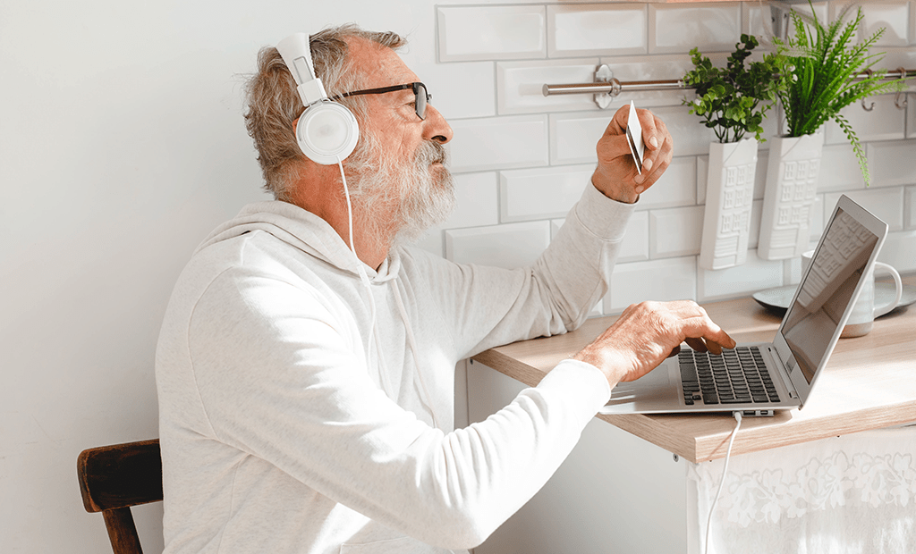 Idoso com fones e óculos usa laptop segurando cartão, ilustrando acessibilidade digital para deficiências sensoriais ou de mobilidade.