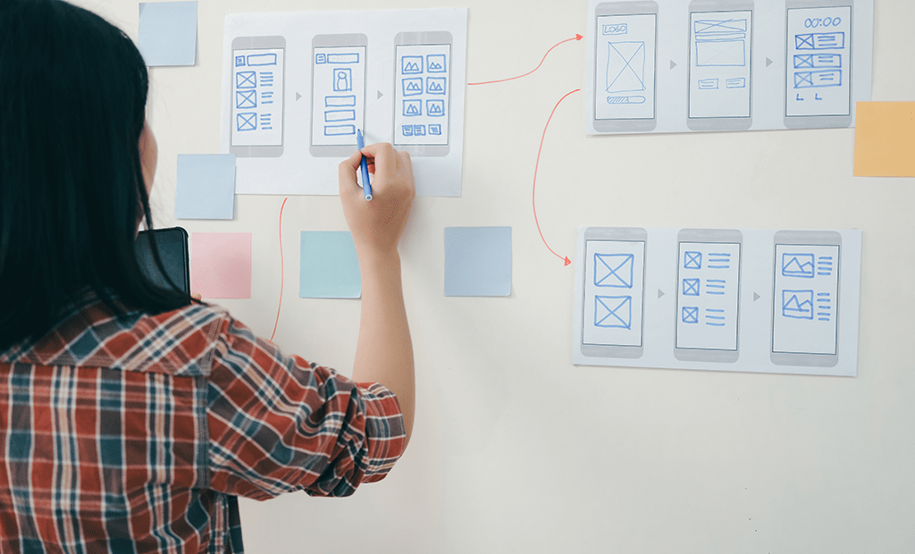 Profissional desenha wireframes de app em papel na parede, mapeando fluxos com setas e post-its para planejar a experiência do usuário.
