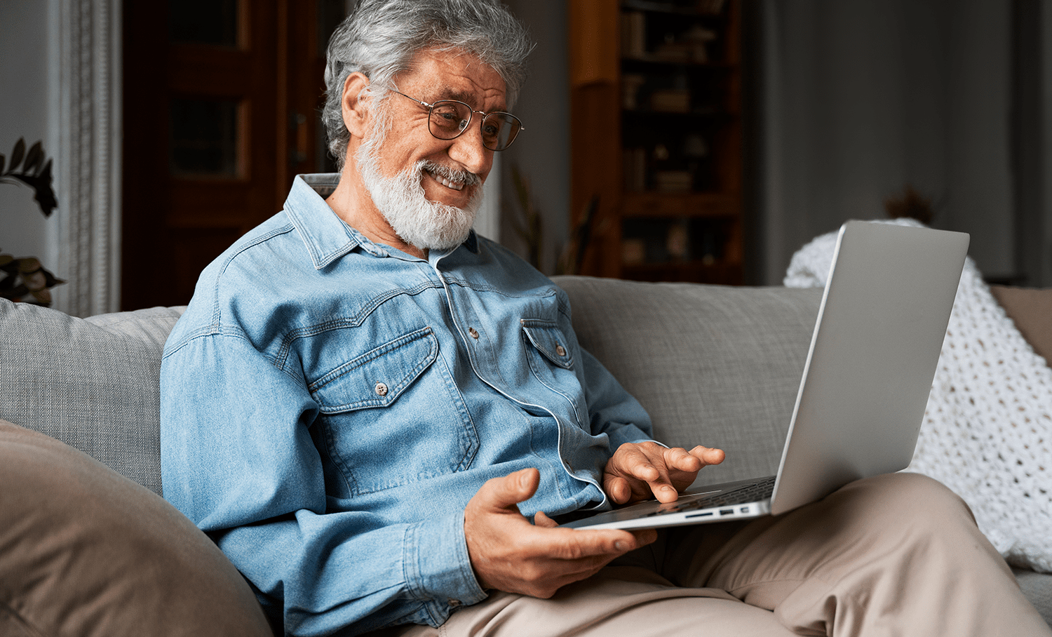 Homem mais velho com barba grisalha e óculos, usando camisa jeans, sorrindo enquanto usa laptop no sofá.