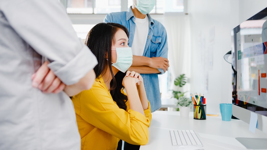Equipe com máscaras analisa gráficos em monitor, ilustrando o contexto de trabalho e acesso à informação durante a pandemia.