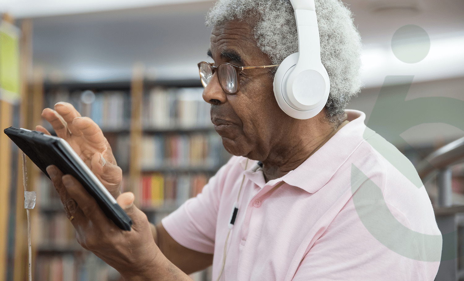 Homem idoso com óculos e fones brancos usa tablet em biblioteca, ilustrando o acesso de seniors à tecnologia digital.