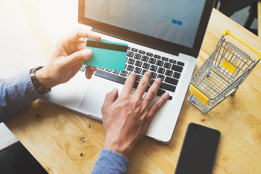 Mãos digitando em laptop com cartão de crédito e carrinho de compras miniatura, representando compras online e acessibilidade no e-commerce.